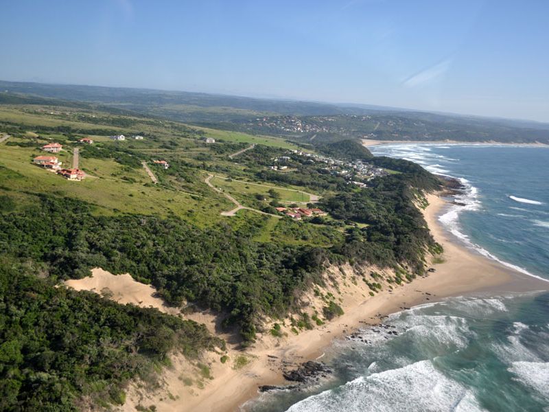 An aerial view of a coastal residential development featuring paved roads, natural vegetation, and direct proximity to the ocean and sandy beach.