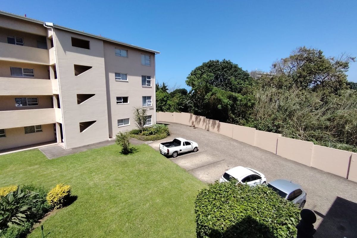 An elevated view of the apartment complex exterior, featuring a paved parking area, landscaped lawn, and perimeter boundary wall.