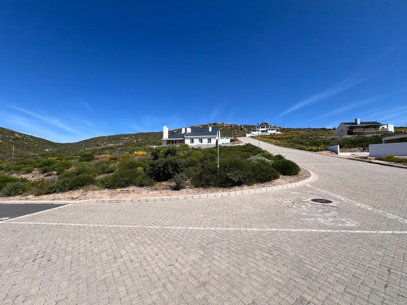 A view of the paved street approach leading up to a residential property set against a hillside landscape.