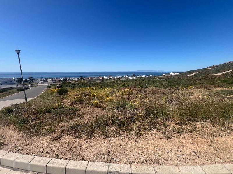 A wide-angle view of a vacant residential plot featuring natural coastal vegetation, paved curbing, and distant ocean views.