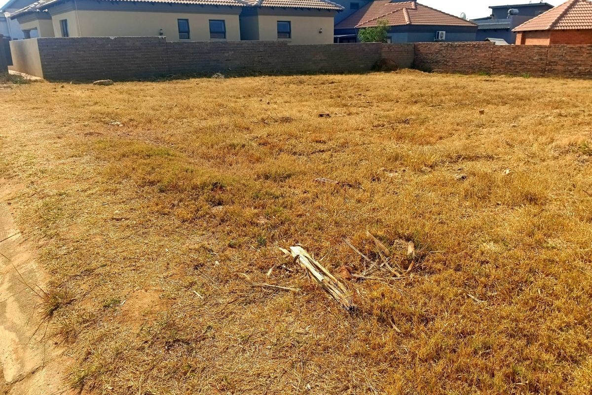 A vacant residential plot featuring dry grass and a perimeter brick boundary wall, situated in a developed residential area.