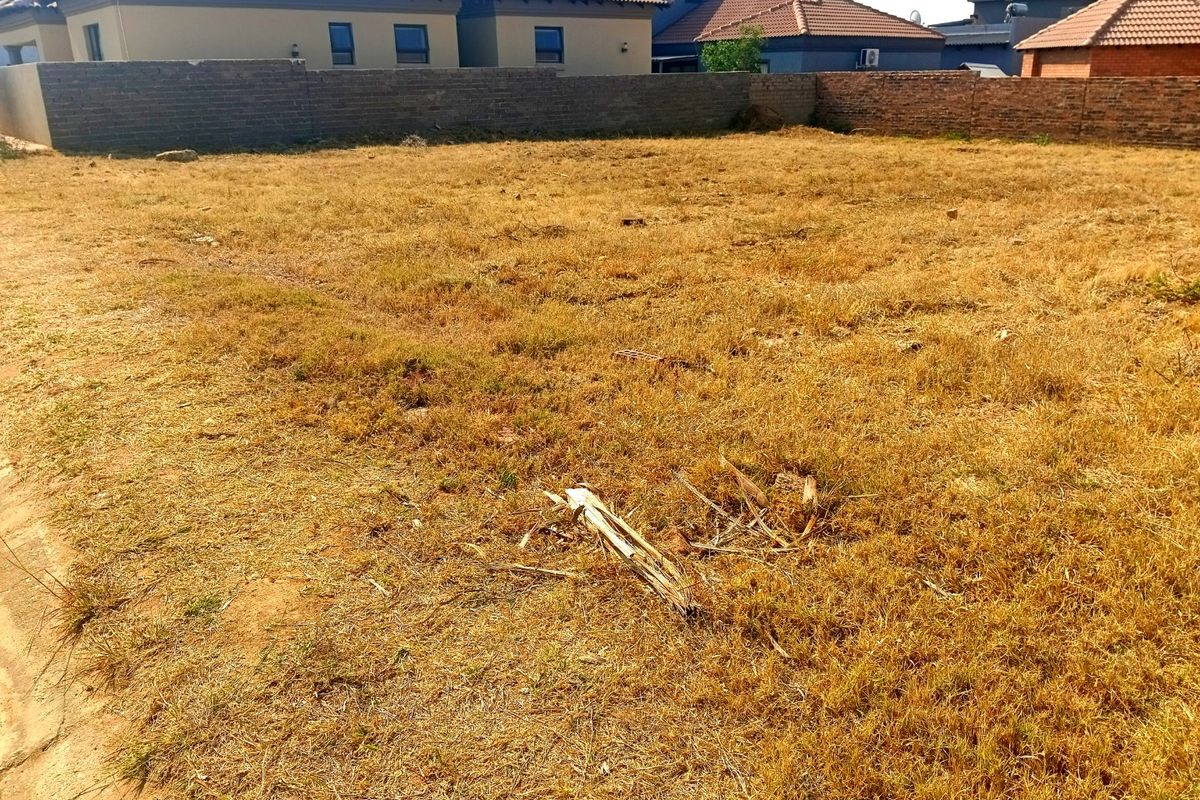 A vacant residential plot featuring dry grass and a perimeter brick boundary wall, situated in a developed neighborhood.