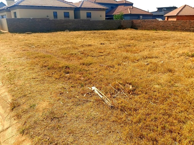 A vacant residential plot featuring dry grass and a perimeter brick boundary wall, situated in a developed neighborhood.