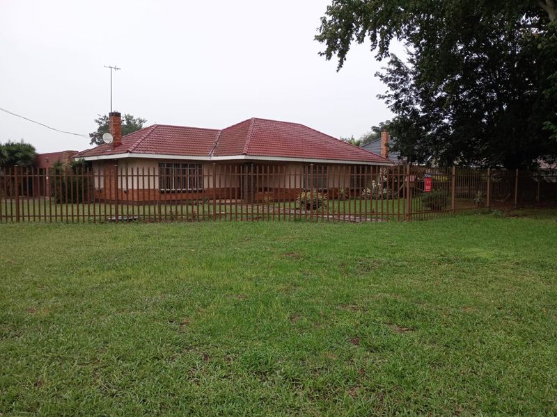 Front view of a single-story brick house with a tiled roof, enclosed by metal perimeter fencing and set on a large grass lawn.