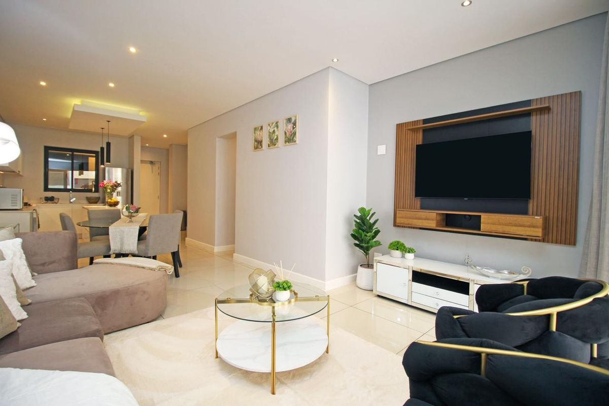 A modern open-plan living and dining area featuring tiled floors, recessed lighting, and a custom built-in media wall, seamlessly connected to a contemporary kitchen.