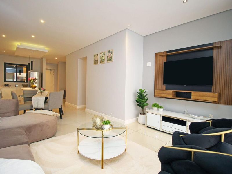 A modern open-plan living and dining area featuring tiled floors, recessed lighting, and a custom built-in media wall, seamlessly connected to a contemporary kitchen.