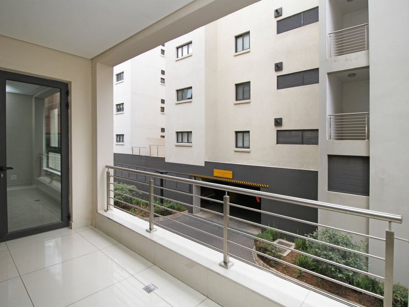 A clean, modern balcony featuring tiled flooring and a stainless steel balustrade, overlooking the apartment complex courtyard and parking entrance.