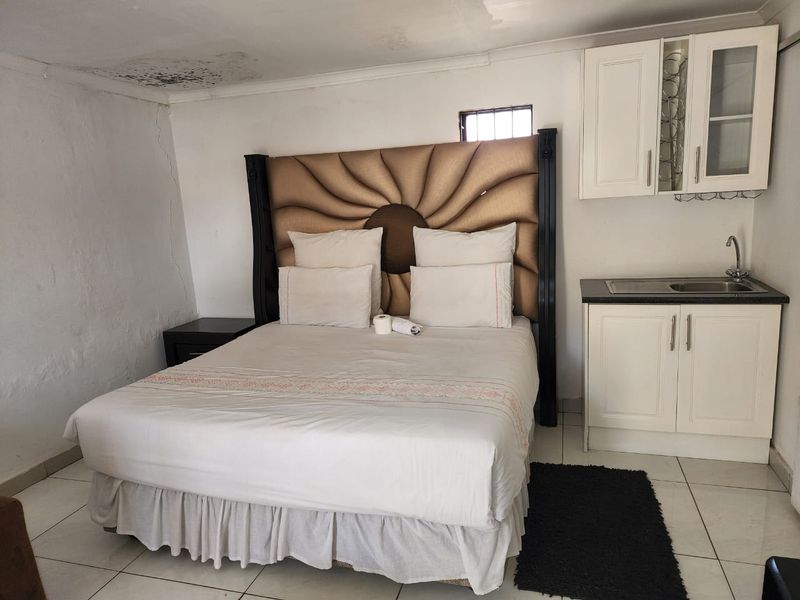 A bedroom featuring a kitchenette area with built-in cabinetry and sink, showing visible signs of ceiling water damage and wall cracking.