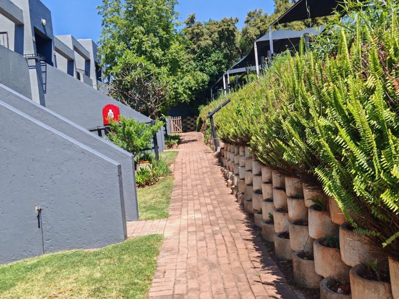 A well-maintained exterior pathway featuring brick paving, a structured retaining wall with integrated planters, and shade sails leading through the townhouse complex grounds.