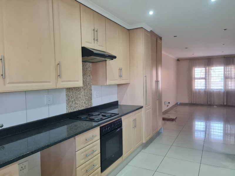 Spacious open-plan kitchen featuring light wood cabinetry, granite countertops, and integrated appliances, flowing seamlessly into a bright, tiled living area.