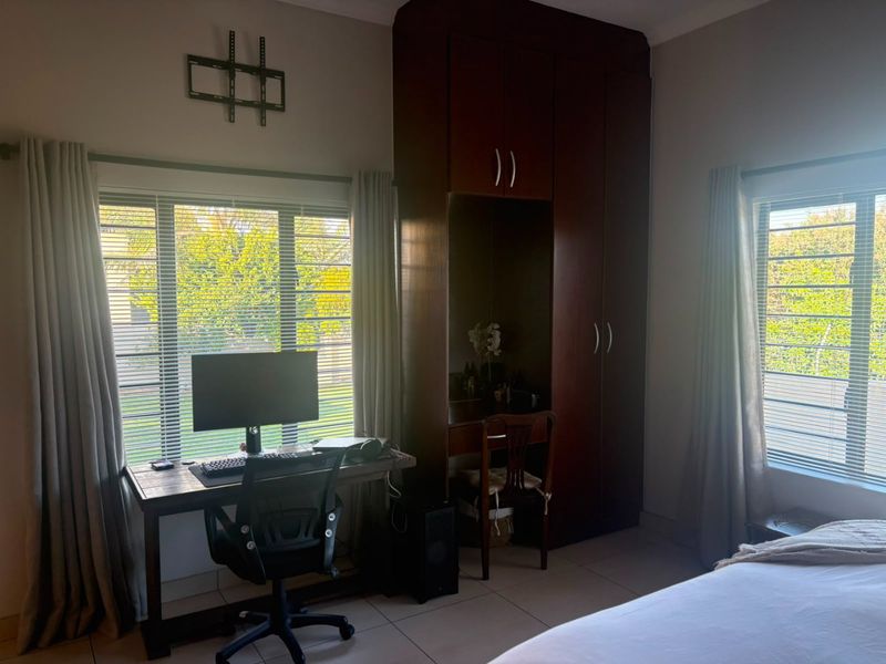 A well-lit bedroom featuring tiled flooring, built-in dark wood cabinetry with a vanity section, and large windows with venetian blinds.