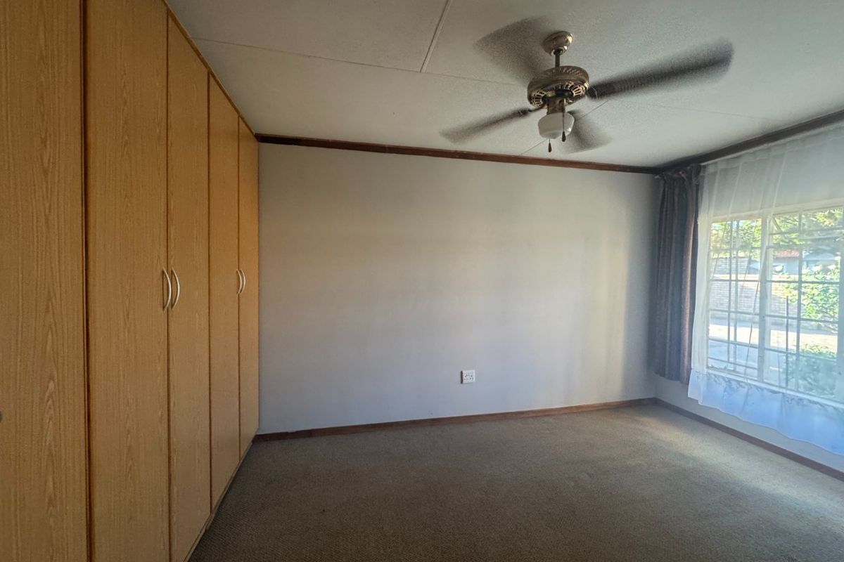 A spacious bedroom featuring built-in wooden cupboards, a ceiling fan, and large windows with security bars, offering a blank canvas for renovation.