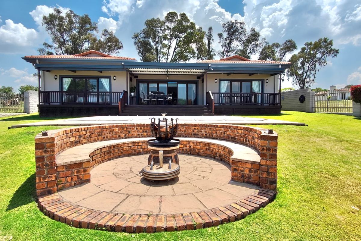 Rear view of the house featuring a raised timber deck, sliding glass doors, and a circular brick fire pit set within a landscaped garden.