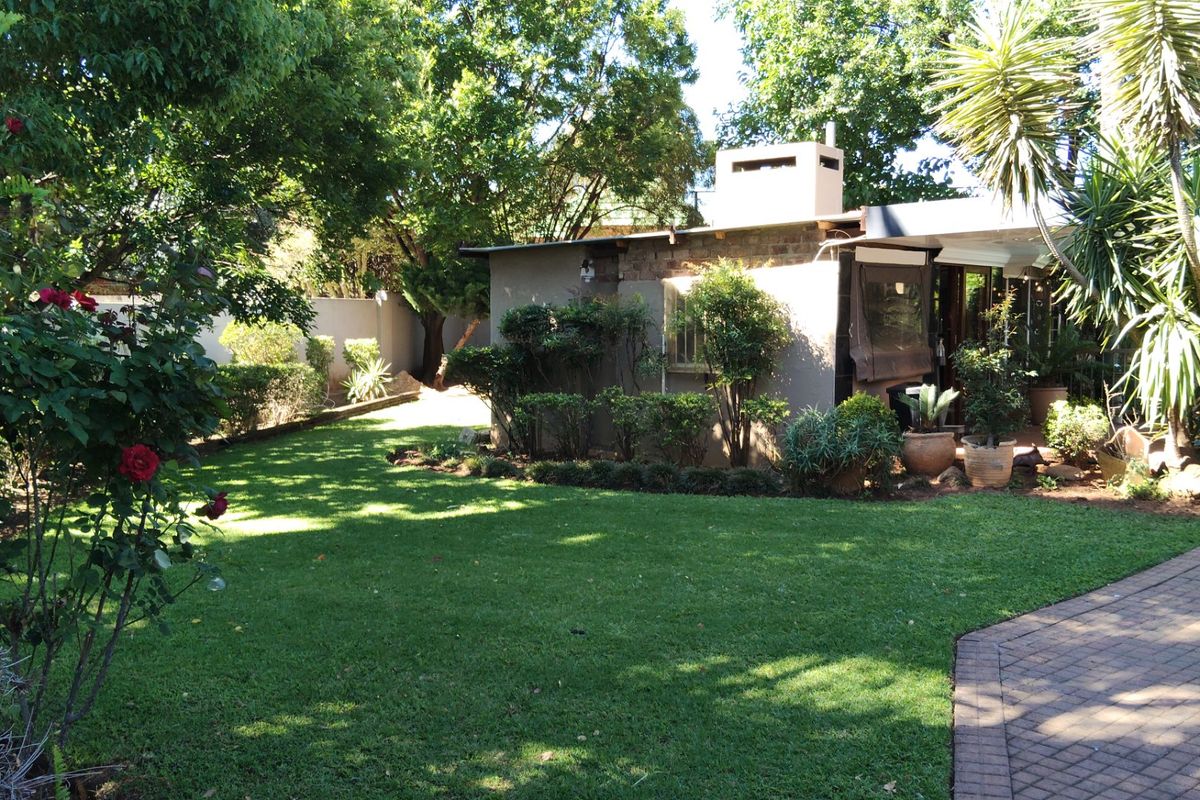 A well-maintained rear garden featuring a lush lawn, mature trees, a brick-paved driveway, and a built-in outdoor fireplace structure.