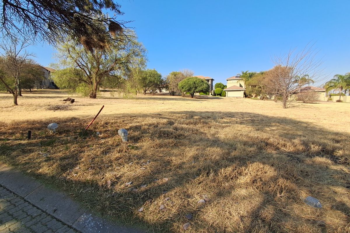 A vacant residential plot featuring dry grass, mature trees, and visible utility connection points, situated in a developed neighborhood.