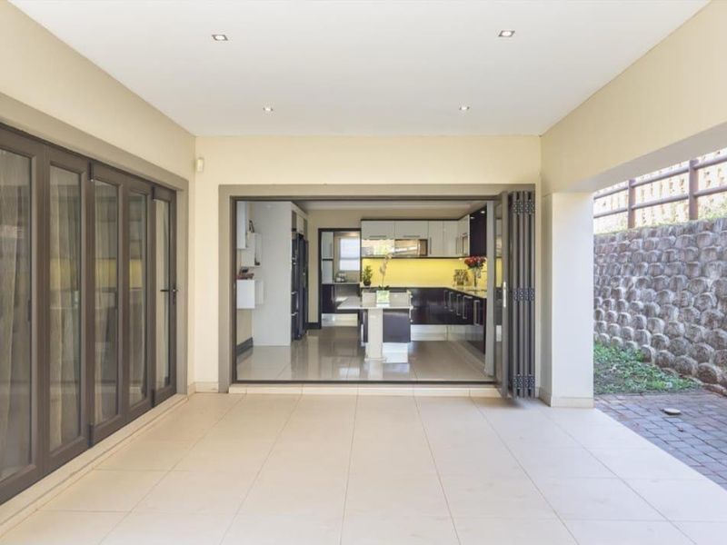 A spacious covered patio featuring tiled flooring and stacking doors that open into a modern kitchen area, adjacent to a stone retaining wall.