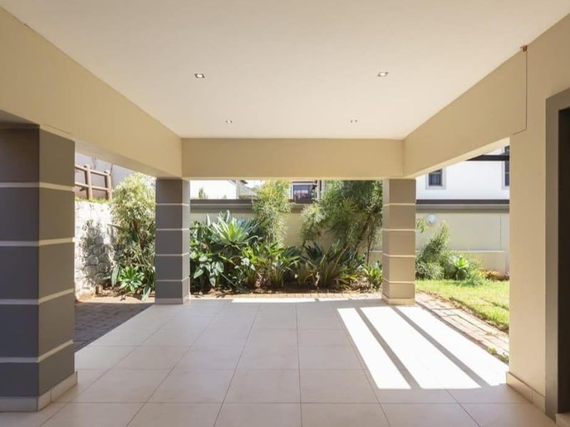 A spacious covered patio featuring tiled flooring, recessed lighting, and support columns, overlooking a landscaped garden area.