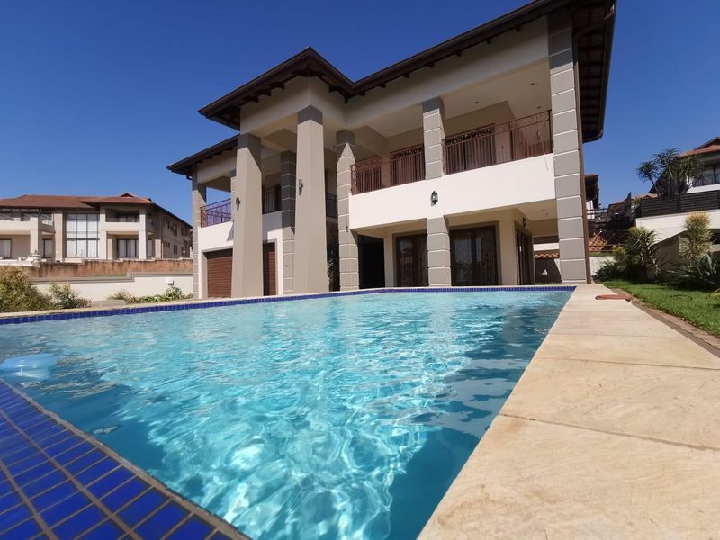 A well-maintained swimming pool area featuring a tiled deck, overlooking a double-story residence with a covered patio and upper-level balcony.
