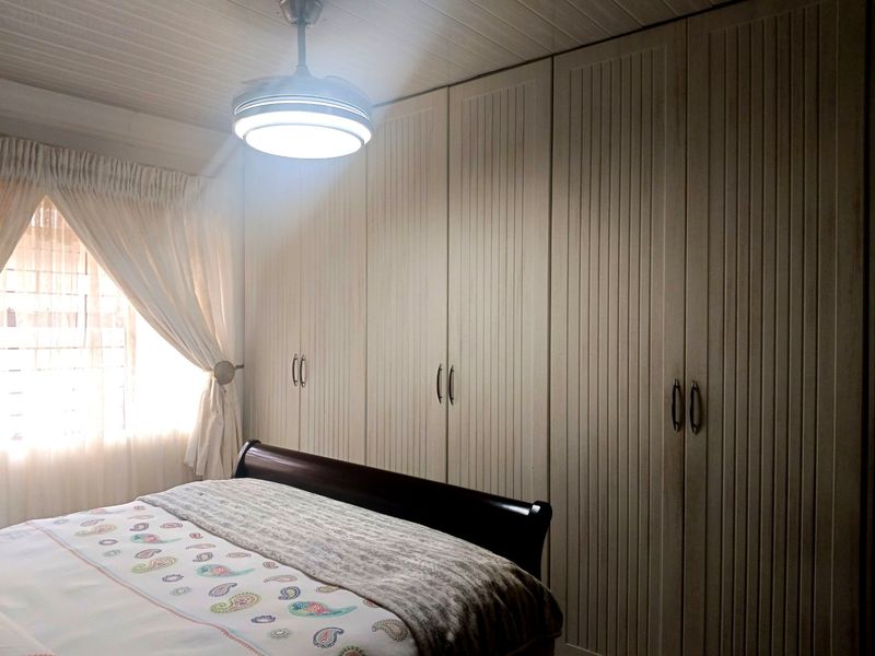 A well-maintained bedroom featuring extensive floor-to-ceiling built-in cupboards, a ceiling fan with light, and classic tongue and groove ceiling paneling.