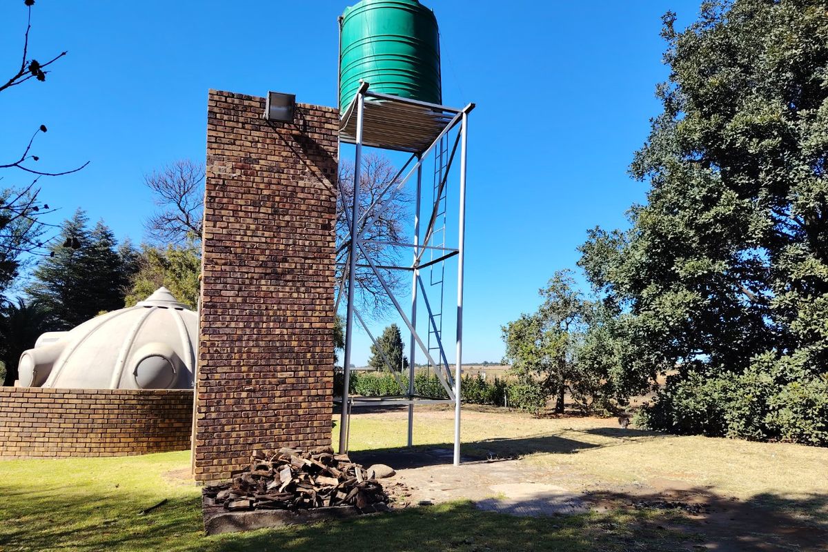 Outdoor area featuring a raised green water storage tank on a steel stand, adjacent to a brick pillar and a dome-shaped structure.