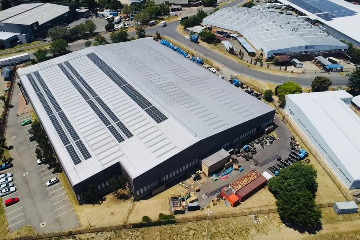 Aerial view of a large industrial warehouse facility featuring a metal roof with integrated solar panels and an adjacent paved parking area.