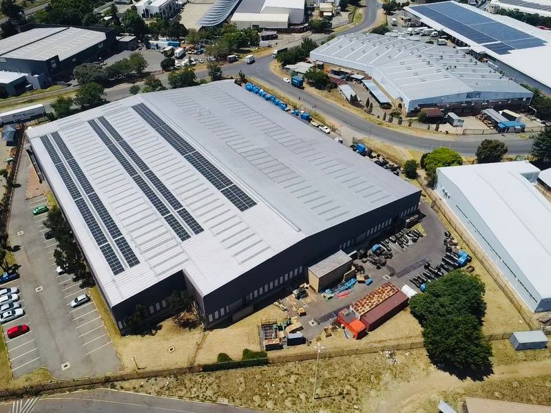 Aerial view of a large industrial warehouse facility featuring a metal roof with integrated solar panels and an adjacent paved parking area.