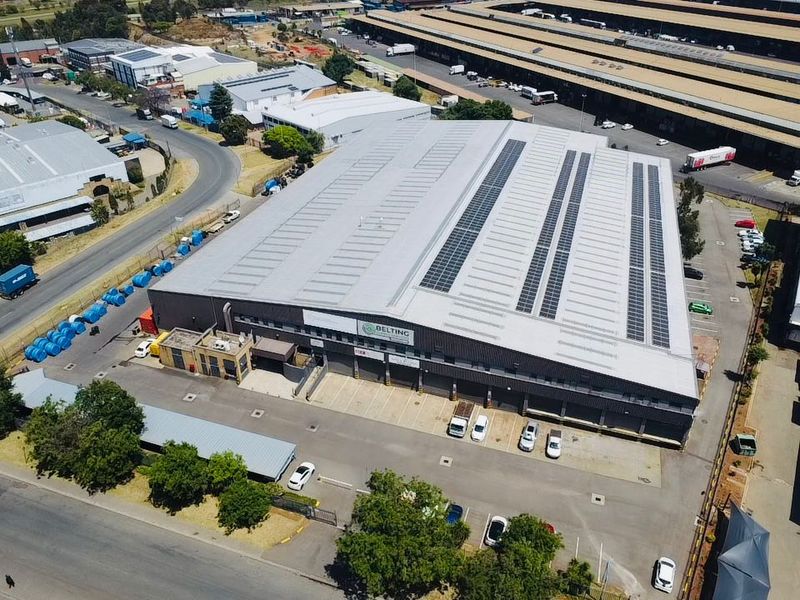Aerial view of a large industrial warehouse facility featuring a metal roof with solar panels, loading bays, and an adjacent asphalt parking area.