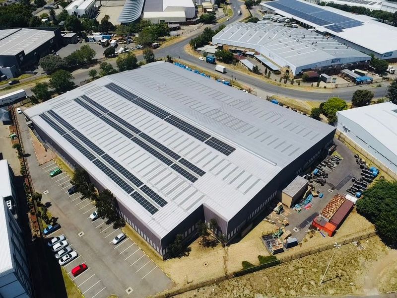 Aerial view of an industrial warehouse facility featuring a large metal roof with integrated solar panels and an adjacent paved parking area.