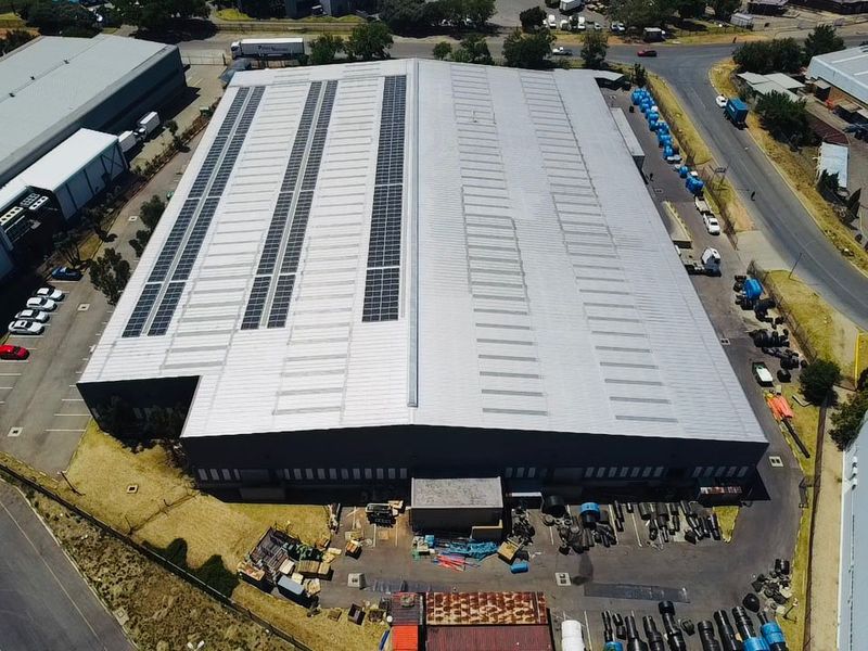 Aerial view of an industrial warehouse facility featuring a large metal roof with integrated solar panels, skylights, and surrounding paved yard space.