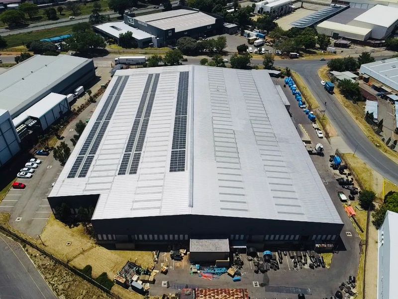 Aerial view of a large industrial warehouse facility featuring a metal roof with integrated solar panels and surrounding paved parking and yard space.