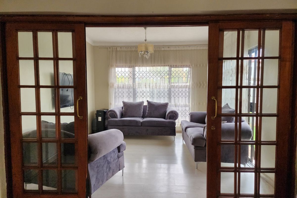 A living room viewed through wooden sliding French doors, featuring laminate flooring and a large window with security bars.