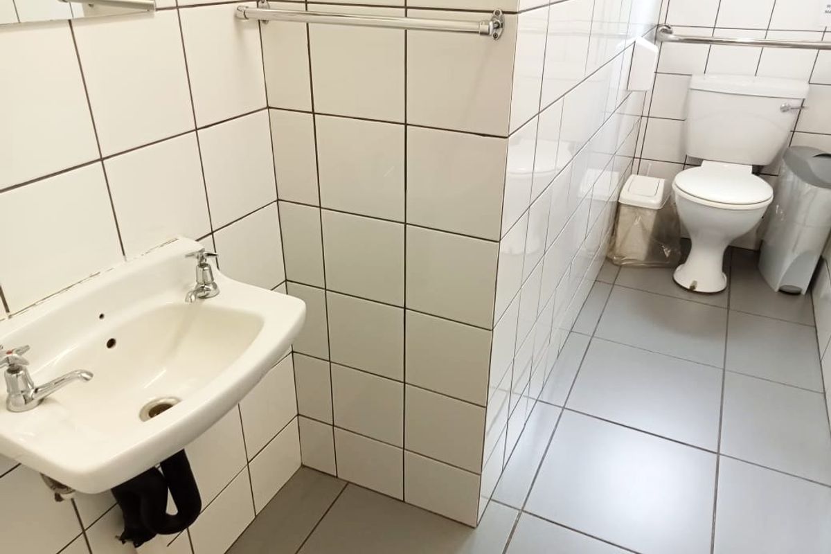 A functional commercial bathroom featuring tiled walls and flooring, equipped with a wall-mounted washbasin, toilet, and safety grab rail.