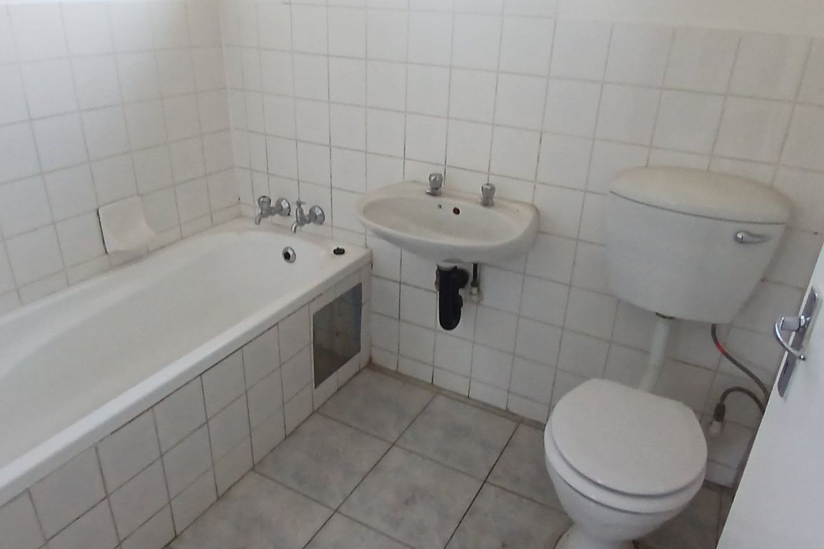A dated bathroom featuring a standard bathtub, wall-mounted basin, and toilet with white ceramic wall and floor tiles.
