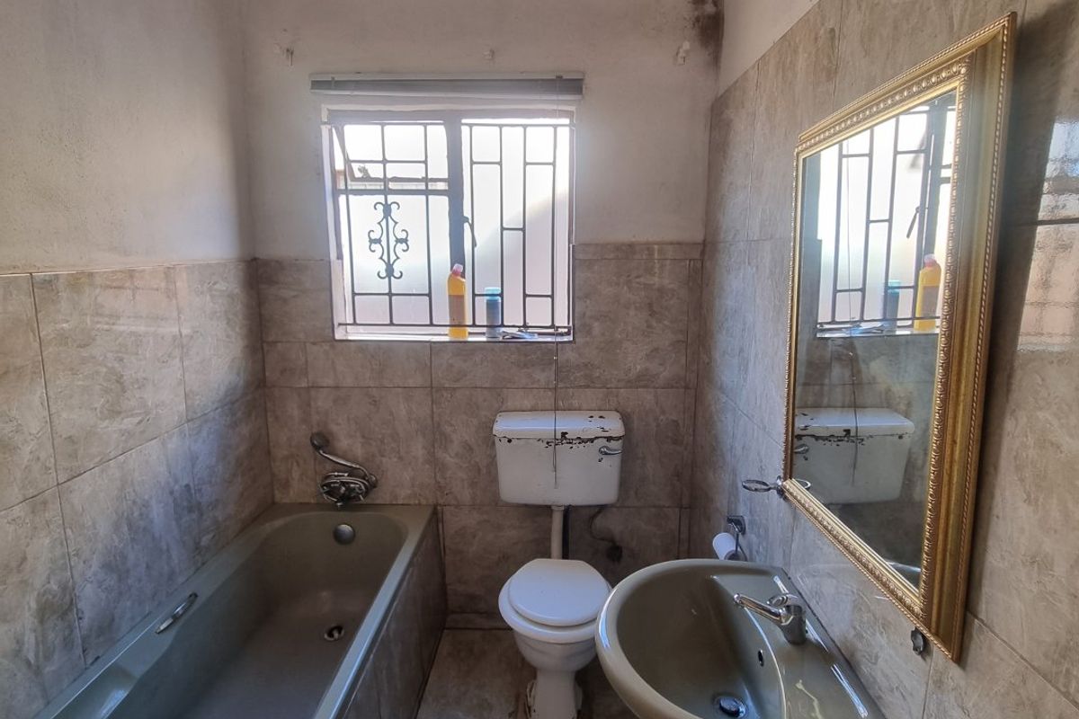 A dated bathroom featuring a bathtub, pedestal basin, and toilet, with tiled finishes and security bars on the window, requiring cosmetic renovation.
