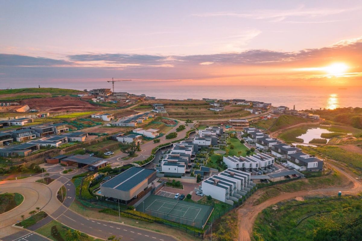 Aerial view of a modern residential estate featuring contemporary housing units, paved infrastructure, and a tennis court, set against a scenic coastal backdrop.