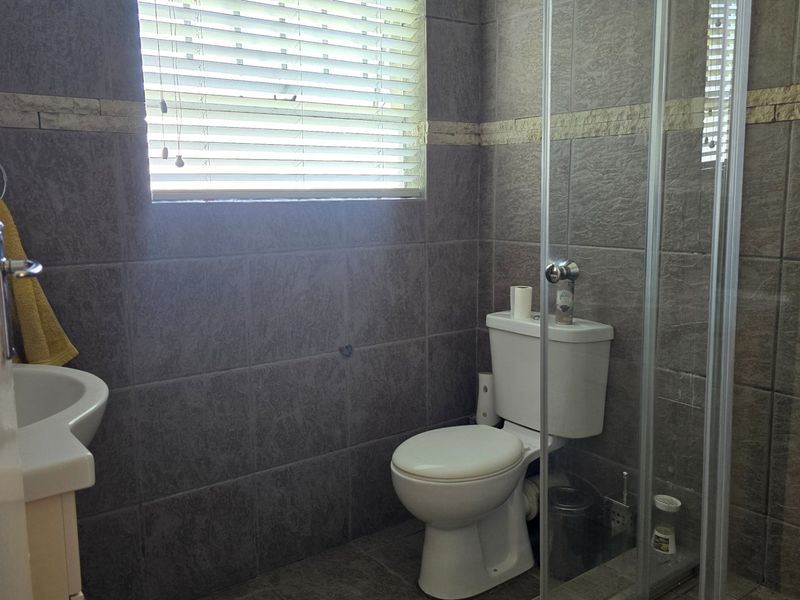 A functional bathroom featuring grey wall tiles, a glass shower enclosure, a pedestal basin, and a toilet with natural light from a window.