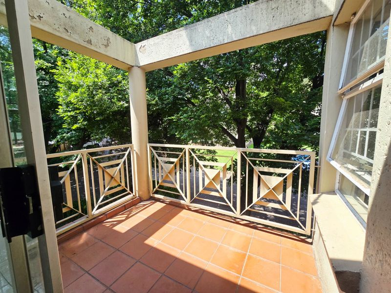 A covered balcony featuring tiled flooring, a decorative metal balustrade, and concrete support pillars overlooking a leafy garden area.