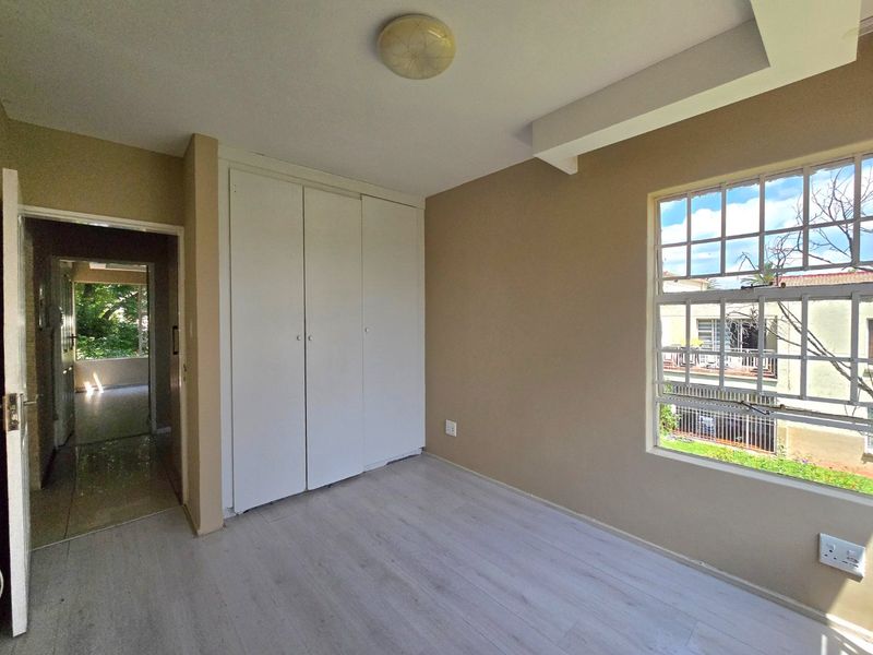 An empty bedroom featuring light-toned laminate flooring, built-in cupboards, and a window with security bars, offering a clean and neutral space.