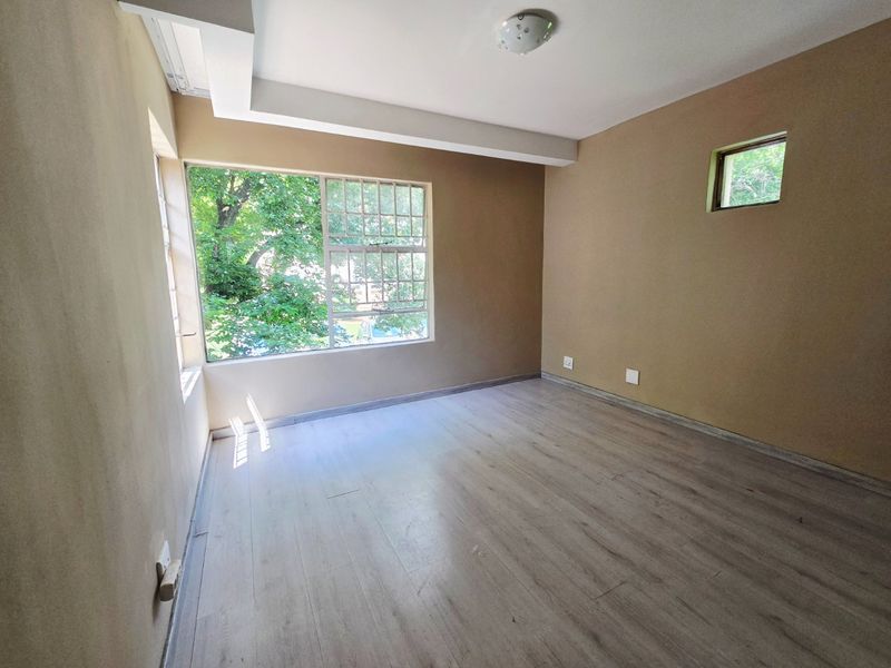 A bright, empty bedroom featuring light-toned laminate flooring, large windows providing natural light, and neutral wall finishes.