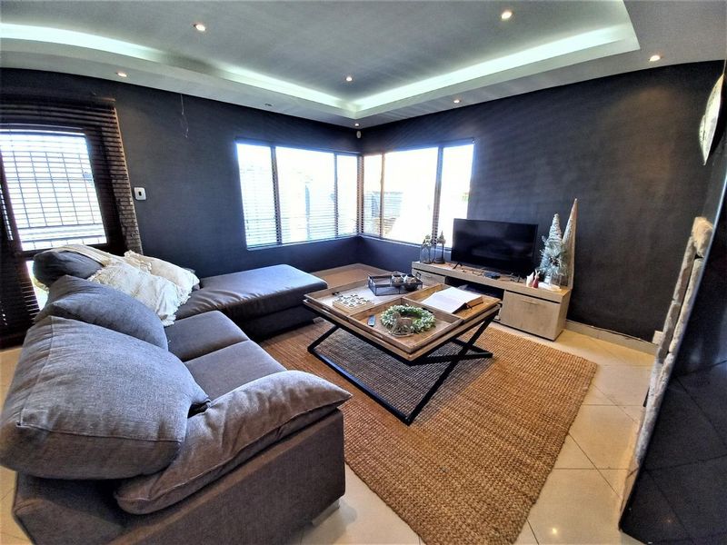 A modern living room featuring dark accent walls, recessed ceiling lighting with cove details, tiled flooring, and large corner windows with venetian blinds.