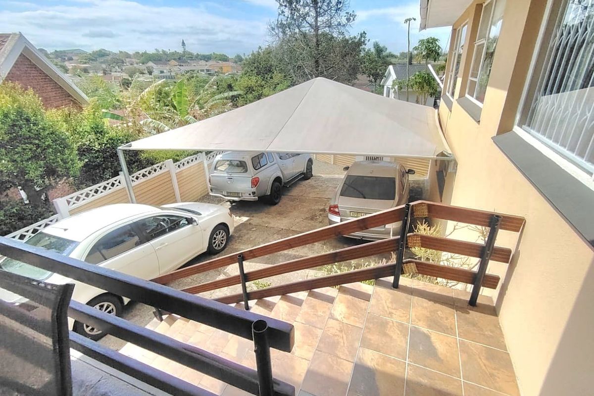 An elevated view of a rear patio area featuring tiled flooring, a timber balustrade, and a large shade sail covering the driveway parking space.