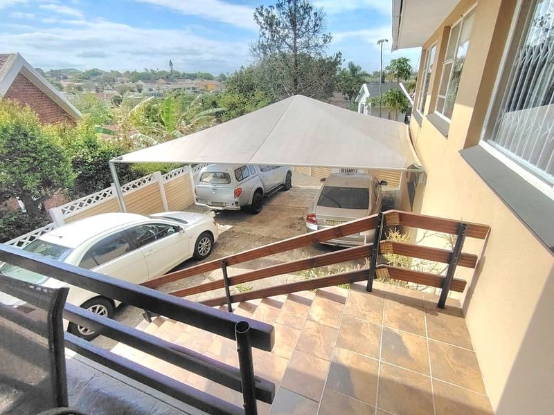 An elevated view of a rear patio area featuring tiled flooring, a timber balustrade, and a large shade sail covering the driveway parking space.