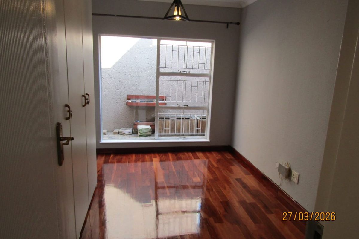 A bedroom featuring polished hardwood flooring, built-in cupboards, and a large window with security bars overlooking a small courtyard area.