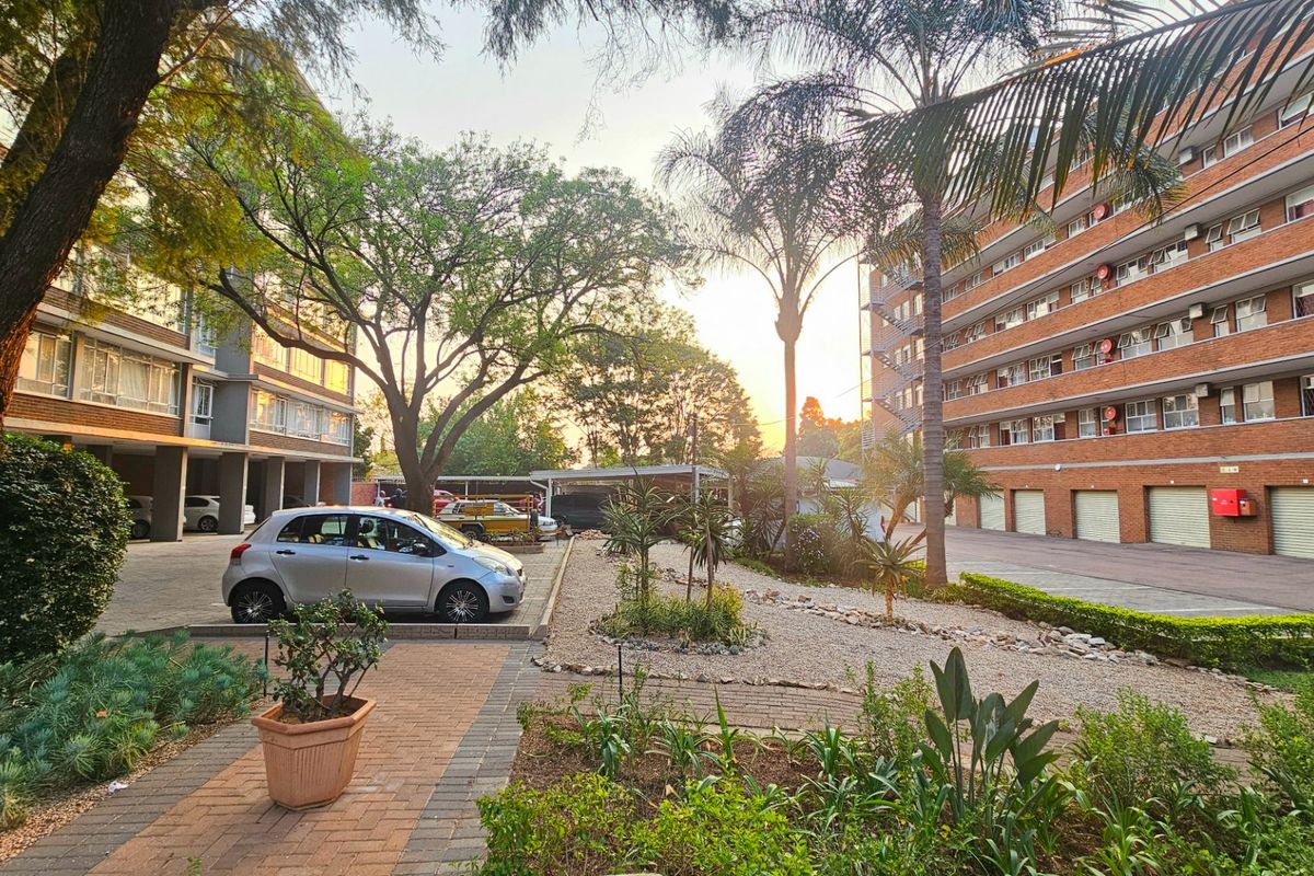 A well-maintained exterior courtyard area featuring paved walkways, landscaped garden beds, mature trees, and access to covered parking facilities within the commercial complex.