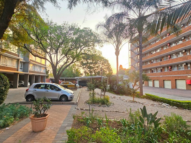A well-maintained exterior courtyard area featuring paved walkways, landscaped garden beds, mature trees, and access to covered parking facilities within the commercial complex.