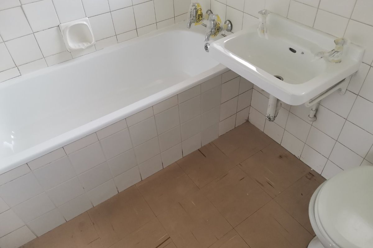 A dated bathroom featuring a standard bathtub, wall-mounted basin, and toilet with original ceramic wall tiling and worn flooring.