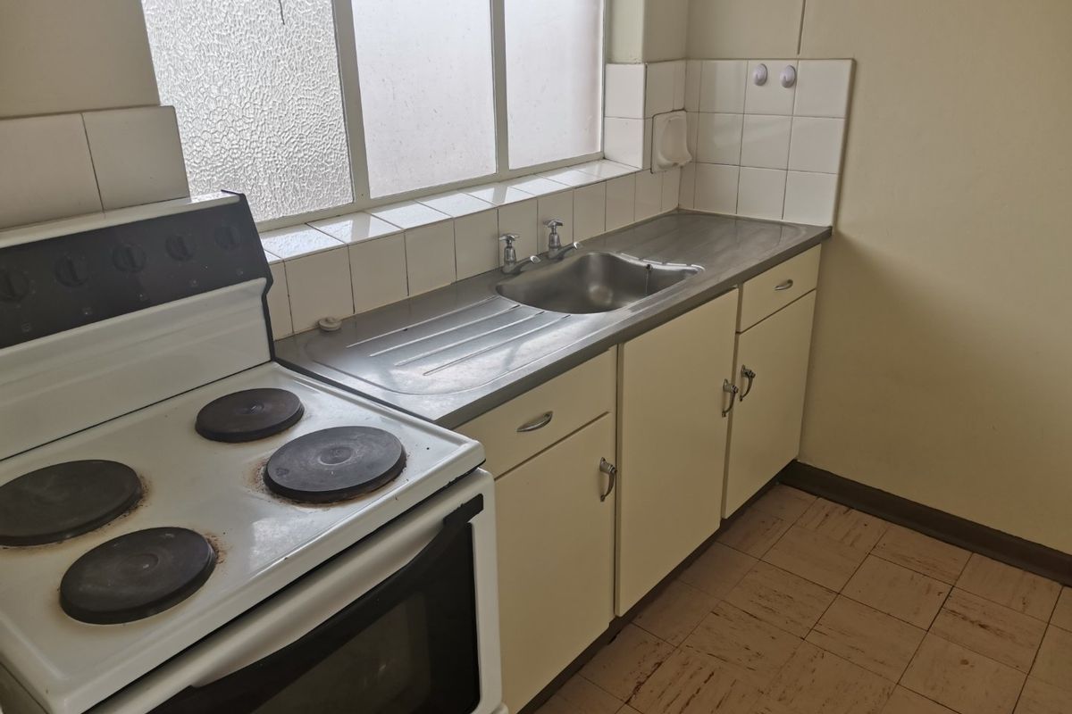 A compact, dated kitchen featuring a stainless steel sink, tiled splashback, and a freestanding electric stove with original cabinetry and vinyl flooring.