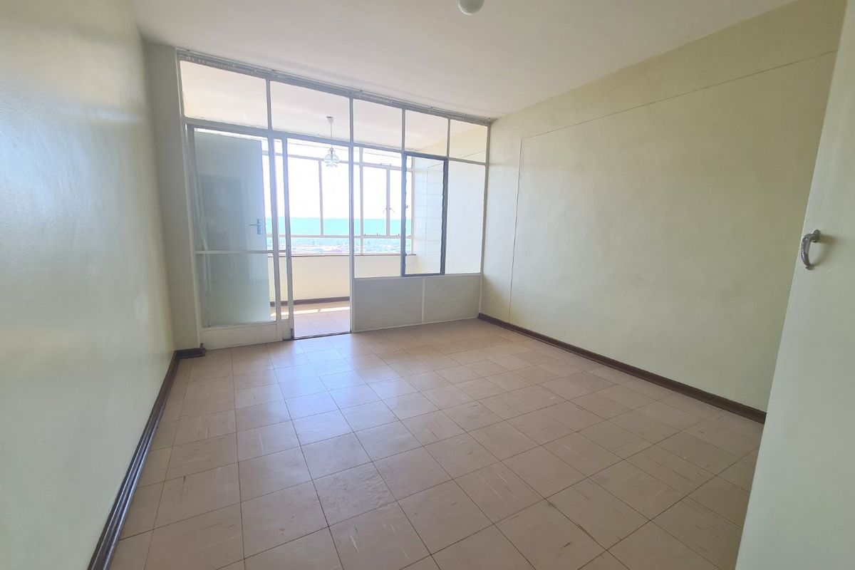 An empty, sunlit living area featuring tiled flooring and a large glass partition leading to an enclosed balcony with exterior views.
