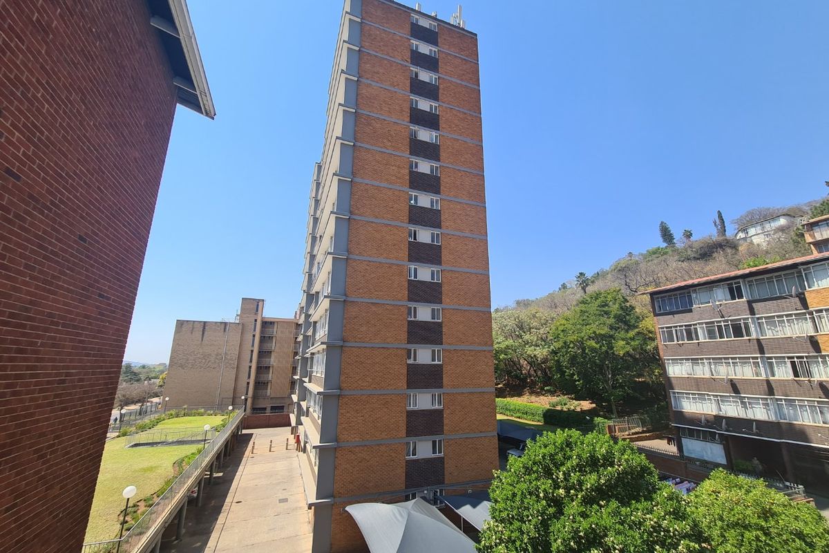 An elevated view of a multi-story brick apartment building with surrounding paved areas, landscaped gardens, and shade structures.