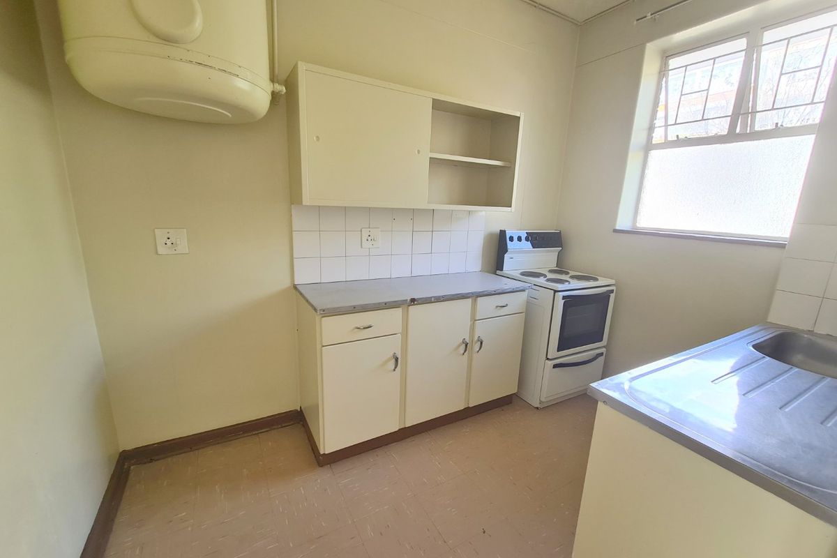 A compact, dated kitchen featuring basic built-in cabinetry, an electric stove, a stainless steel sink, and a wall-mounted electric water heater.
